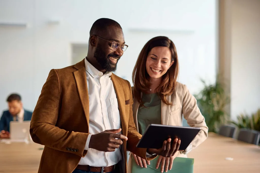 Two happy business colleagues engaging with a digital tablet and discussing various projects while standing in a bright, modern office environment, showcasing teamwork and collaboration