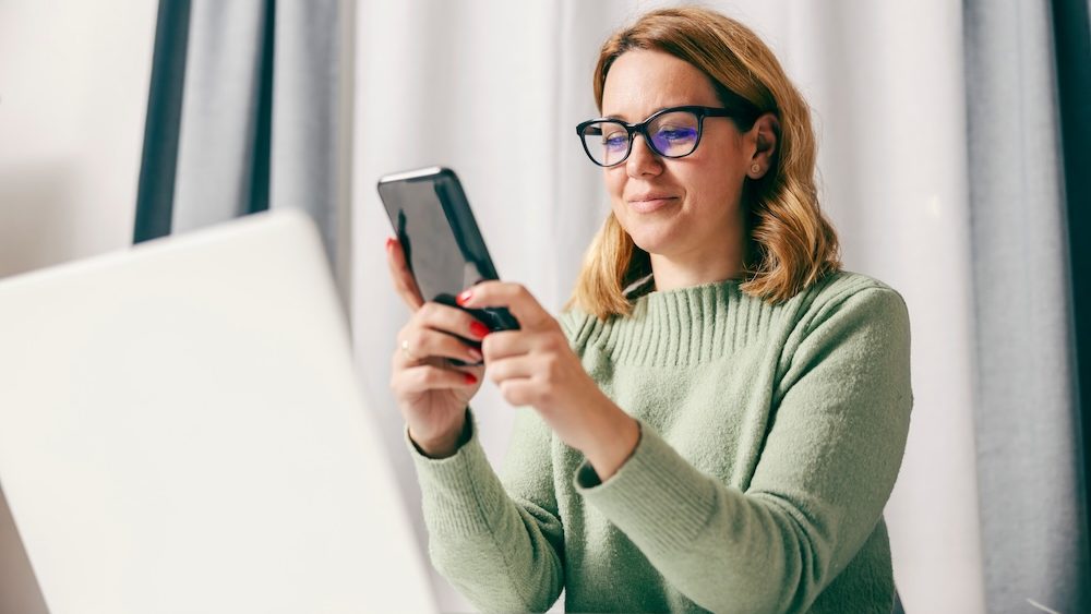 A happy entrepreneur using smart phone while working online at her home office.